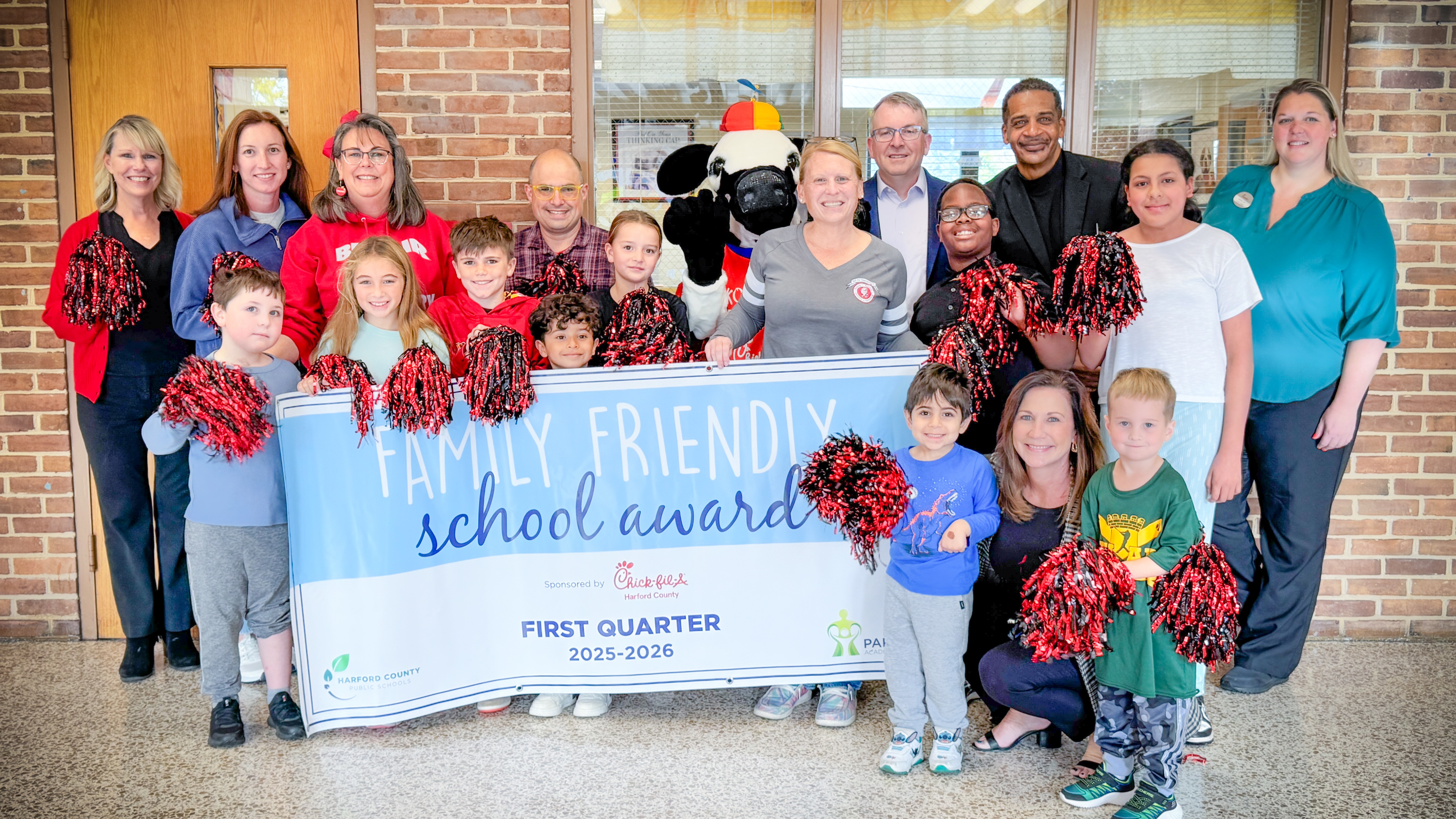 School system leaders, students, and Chick-fil-A mascot hold up First Quarter Family Friendly School Award banner.