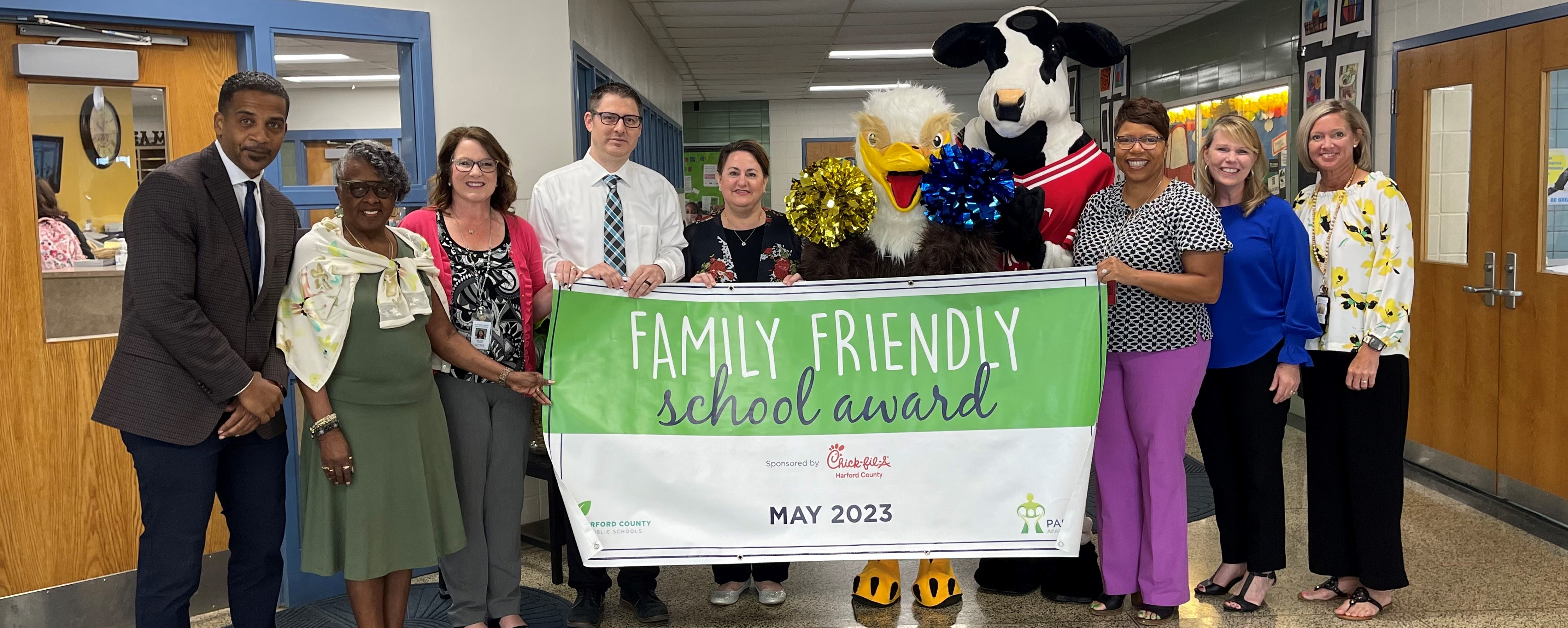 School system leaders, school staff, and mascots hold up May 2023 Family Friendly School banner
