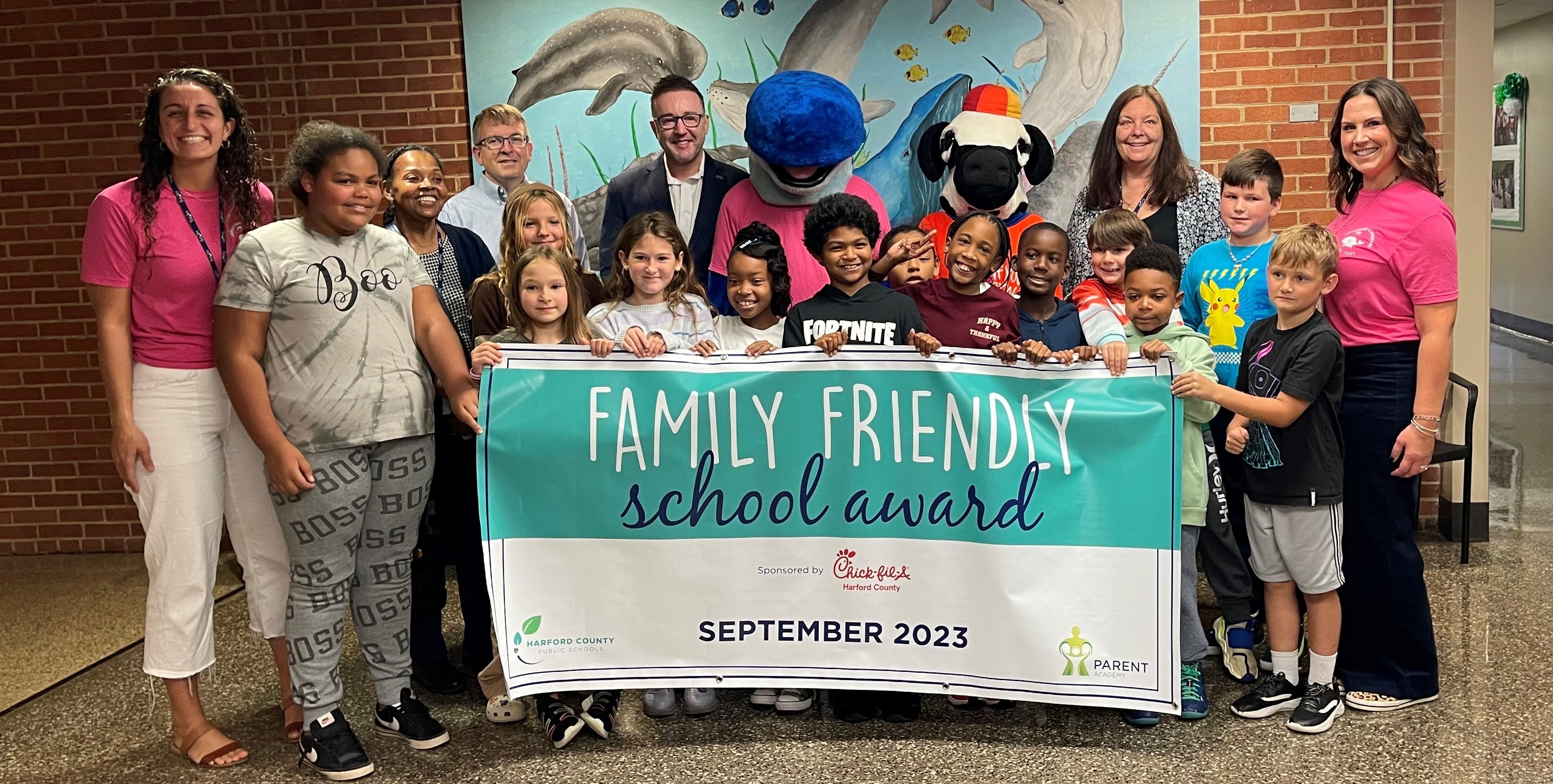 School system leaders, school staff, students, and mascots hold up September 2023 Family Friendly School banner