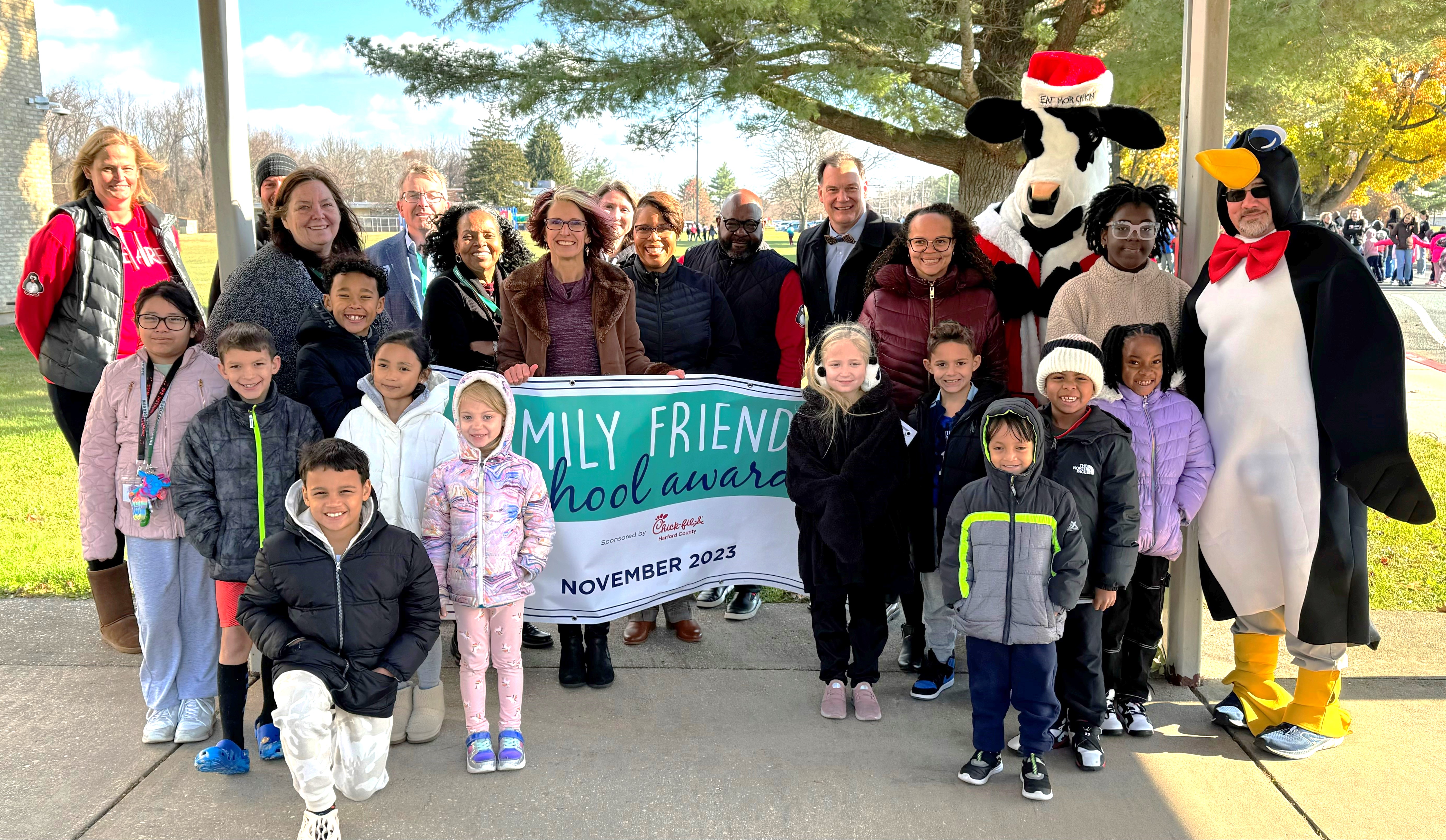 School system leaders, school staff, students, and mascots hold up November 2023 Family Friendly School banner.
