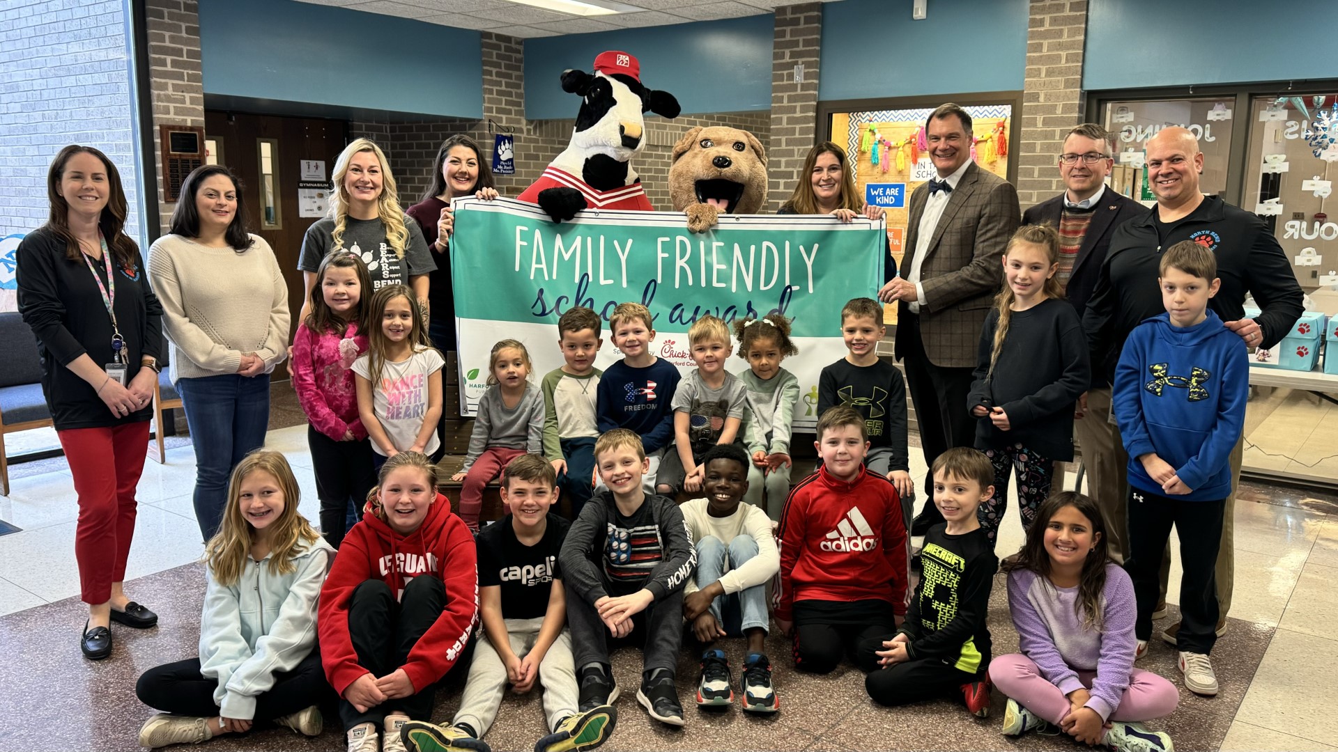 School system leaders, school staff, students, and mascots hold up January 2024 Family Friendly School Award banner.