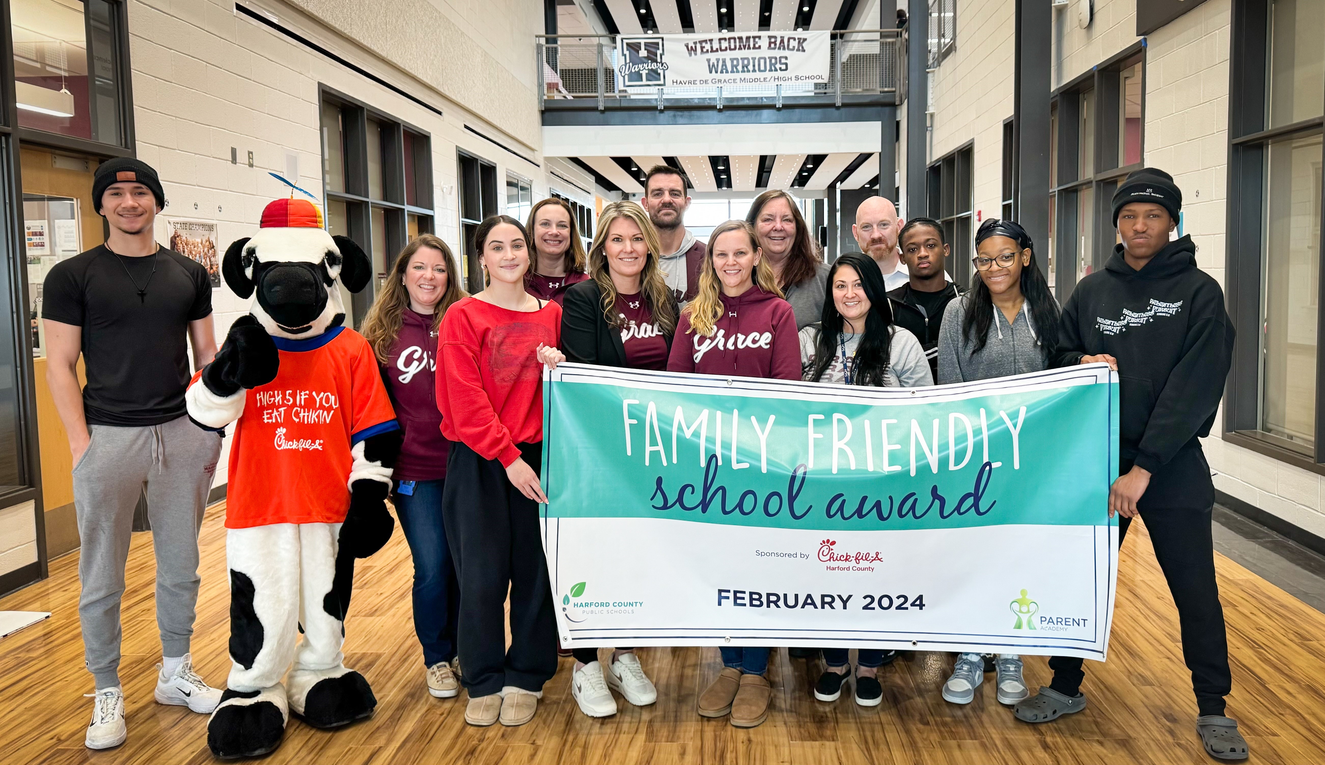 School system leaders, school staff, students, and Chick-fil-A mascot hold up February 2024 Family Friendly School Award banner.