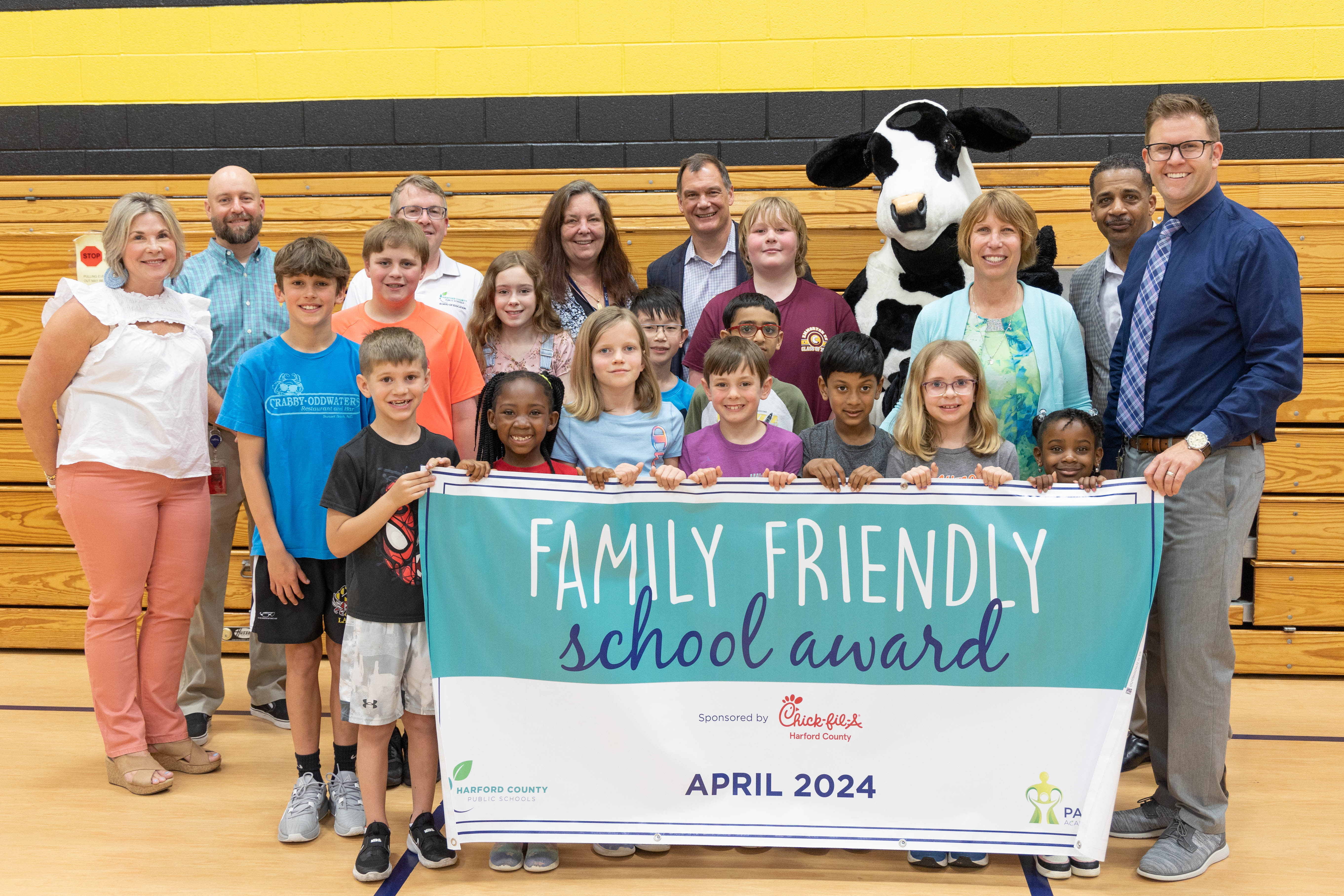 School system leaders, students, and Chick-fil-A mascot hold up April 2024 Family Friendly School Award banner.