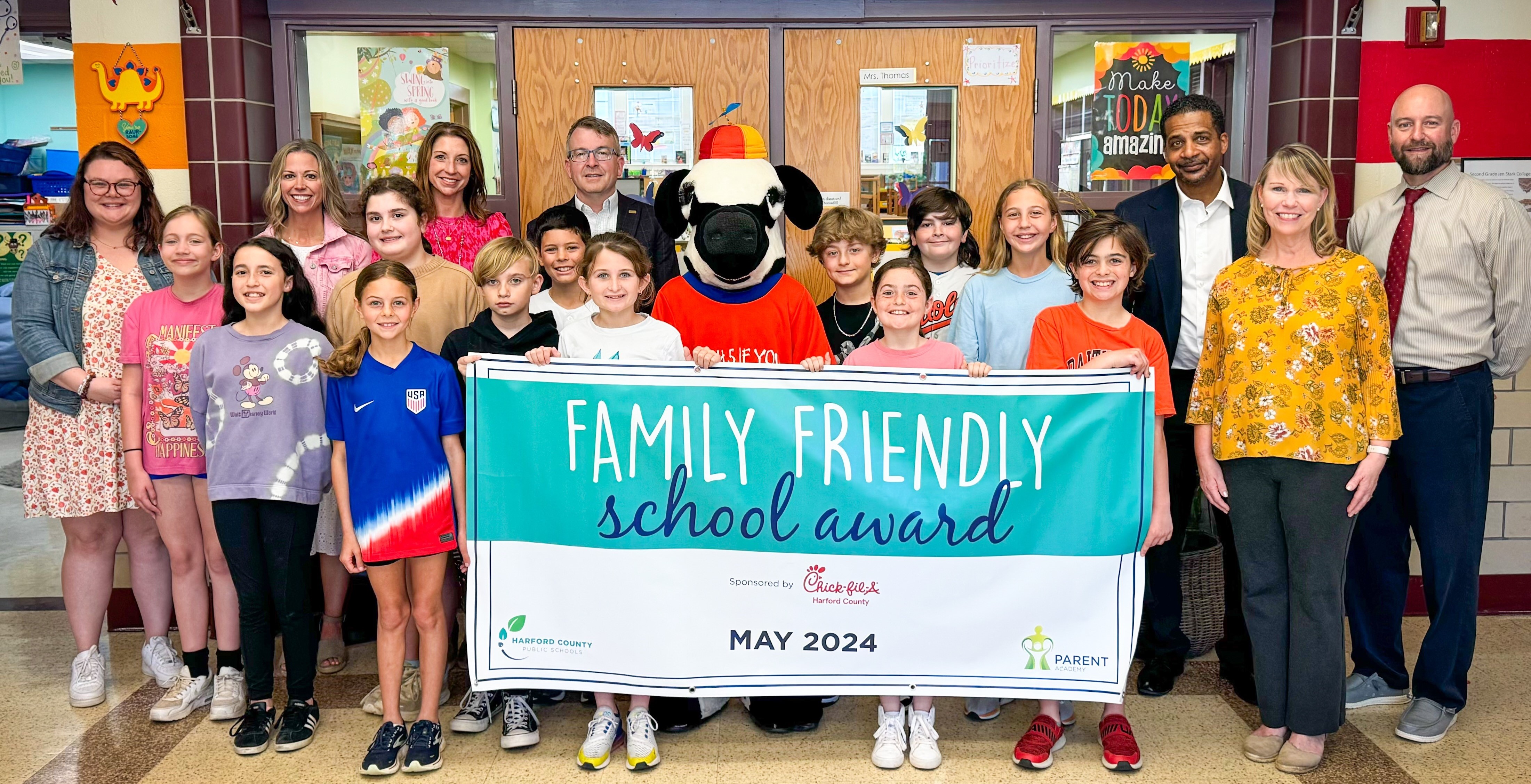 School system leaders, students, and Chick-fil-A mascot hold up May 2024 Family Friendly School Award banner.